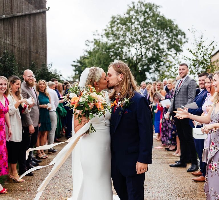 Bride holding a colourful wedding bouquet sharing a kiss with the groom at the end of their confetti exit 