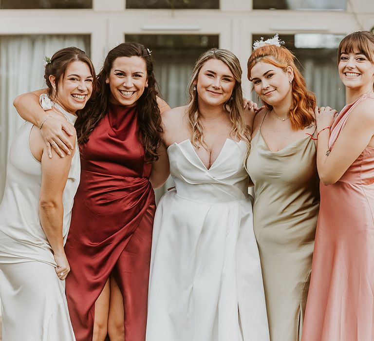 Bridal party in mismatched satin bridesmaid dresses smiling with the bride in Suzanne Neville wedding dress 