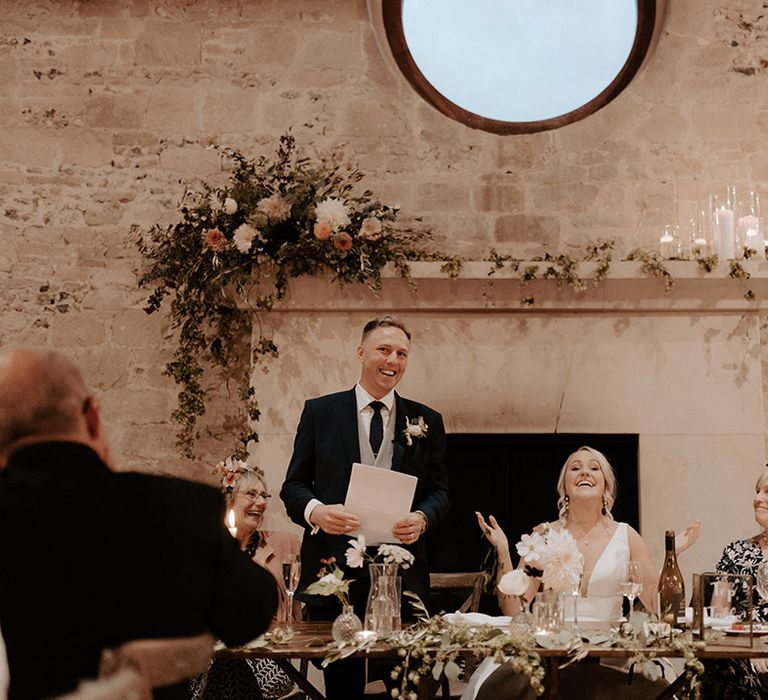 Groom stands up to perform his wedding speech with the bride smiling happily 