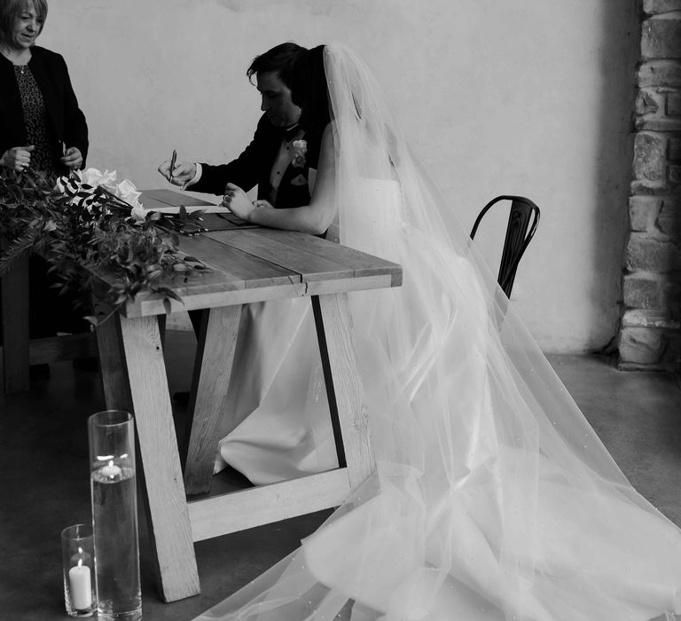 Bride in long personalised veil signing the marriage certificate with groom in black tuxedo