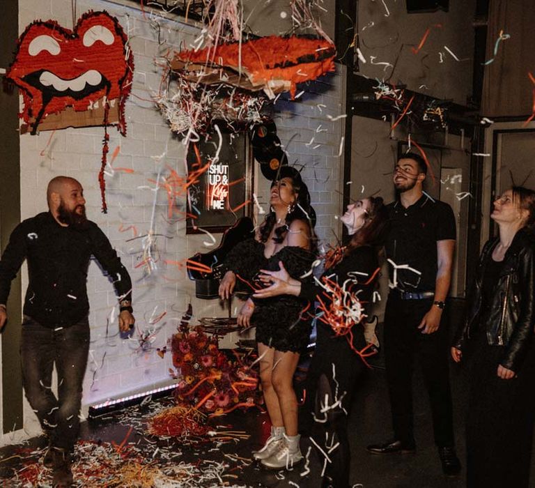 Bride in black ruffle off shoulder reception dress and red drip effect sunglasses with groom in black shirt and black drip effect sunglasses standing with wedding guests batting the Rolling Stones wedding piñata 