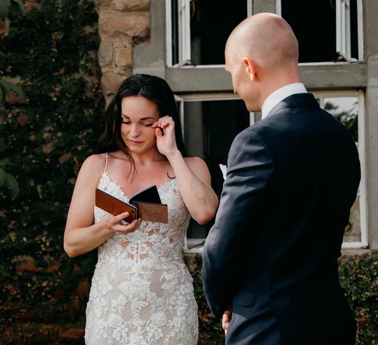 Bride in lace sleeveless wedding dress with puddle train reading from her personalised vow book with groom in classic black grooms suit 