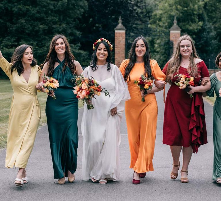 Bride in long sleeve mesh overlay wedding dress and colourful bridal flower crown walking on the grounds of Templars Barn with bridesmaids in satin mismatched bridesmaid dresses