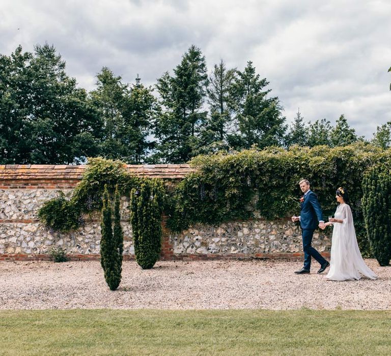 Groom in classic navy grooms suit with black tie, grey waistcoat and colourful rose boutonniere walking with bride in long sleeve mesh overlay wedding dress and colourful bridal flower crown on the grounds of Templars Barn wedding venue