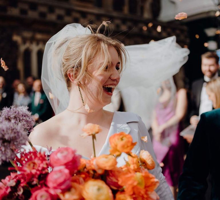 Bride in asymmetrical off shoulder long sleeve satin wedding dress with cathedral length personalised veil holding colourful bridal bouquet with pink garden roses, purple giant alliums, dark red poppies and foliage at Fabrica Brighton wedding doing colourful petal confetti exit