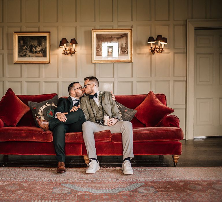 The two grooms sit on their red velvet sofa at the wedding venue and share a kiss 