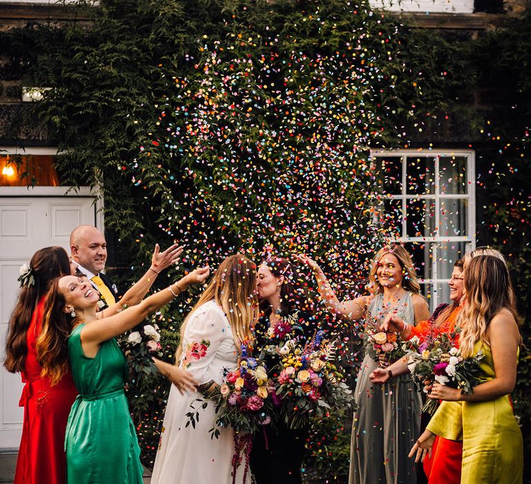 Bridesmaids in mismatched bridesmaid dresses in bold colours throw confetti over the two brides 