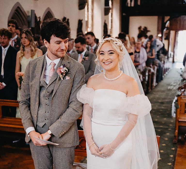 Groom in three piece grey tweed suit standing with the bride in an off the shoulder puff sleeve wedding dress 