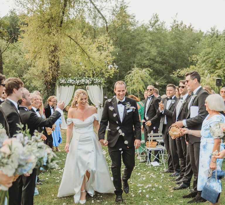 Wedding guests lining the aisle throw white petal confetti over the bride and groom 