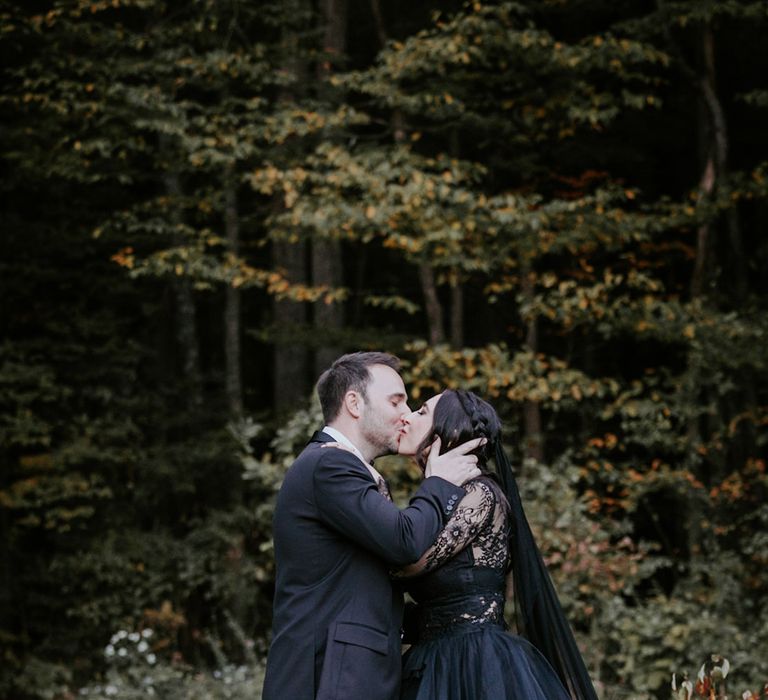 Bride in bespoke black wedding dress with long sleeves kissing the groom in a black tuxedo 