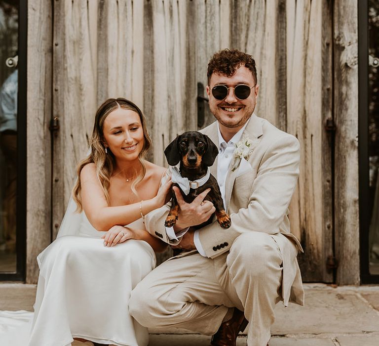 The bride and groom pose with their pet dog wearing a silver bow collar for the wedding 