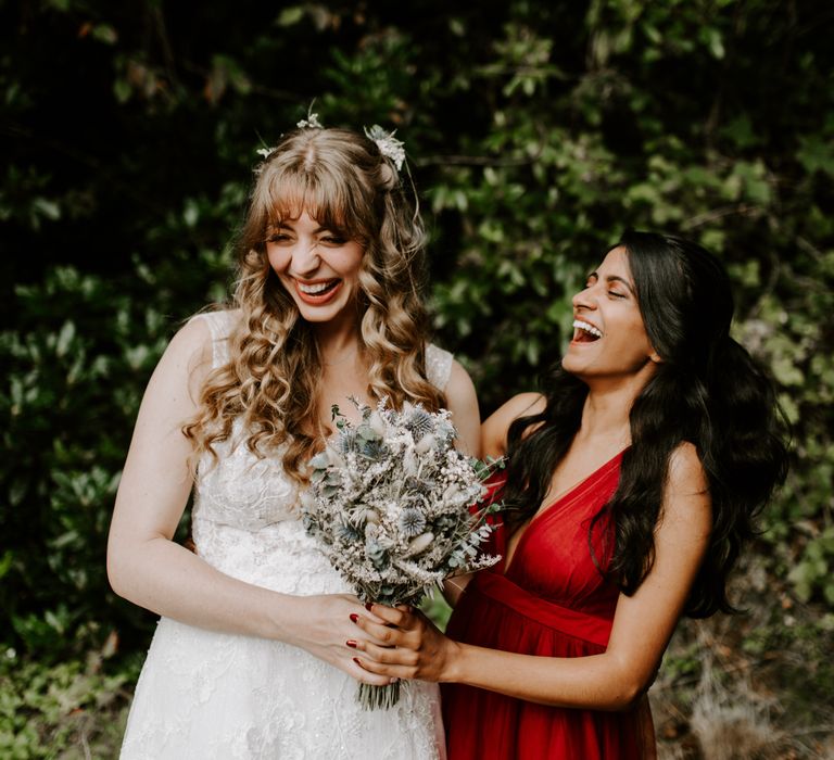 Bride laughing with bridesmaid in low cut red dress with a dried flower bouquet of pampas grass and thistle and green foliage