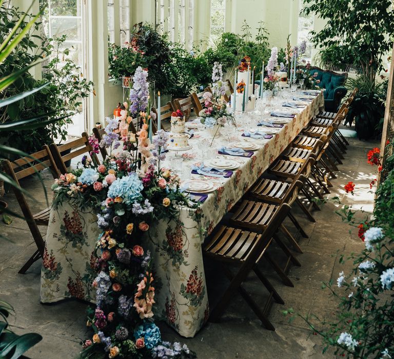 Floral tablecloth with deep purple napkins and blue ribbons and tapered blue candles with three wedding cakes and tall centrepieces in conservatory 