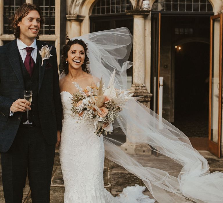 Groom in a navy check suit and burgundy tie standing with his bride in a strapless lace wedding dress sipping champagne 