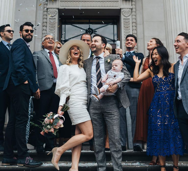 Confetti moment with bride and groom holding their baby celebrating with close family and friends at intimate London wedding with short wedding dress