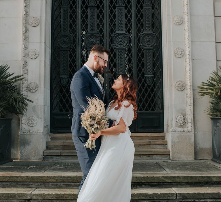 Bride in white dress and celestial headband holds dried flower bouquet whilst hugging groom in navy suit on steps