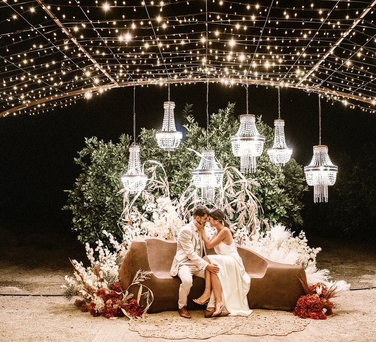 Bride and groom sitting at an outdoor seating area with fairy light canopy and chandelier light installation 