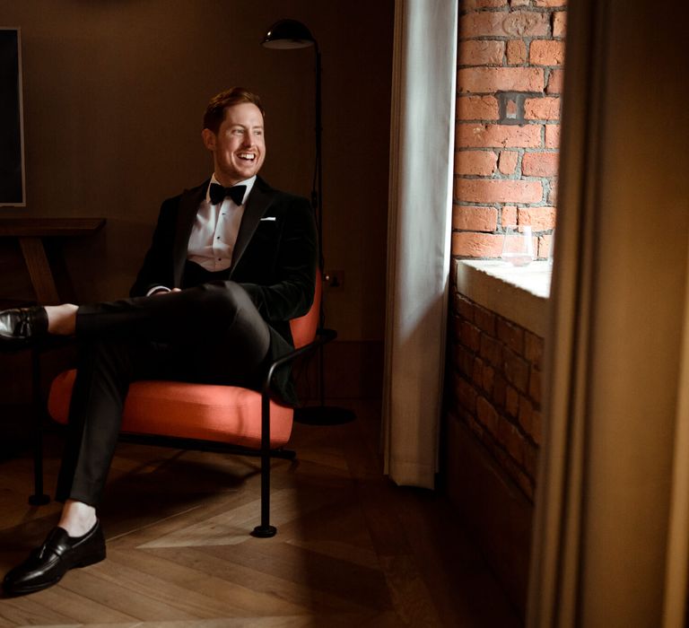 Groom wearing three piece suit and bow tie sat down laughing before his wedding