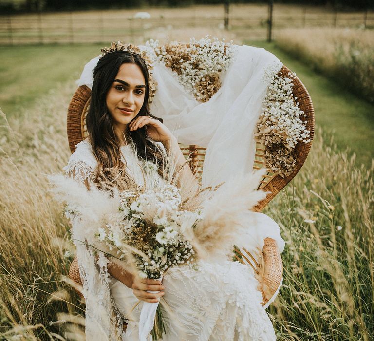 Boho bride sitting in a field in a peacock chair in a lace Julita London wedding dress holding a dried flower bouquet 