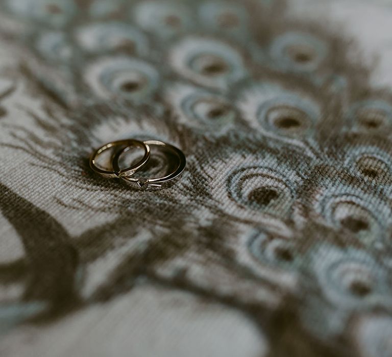 Two wedding rings lying on grey woven fabric with peacock feather pattern