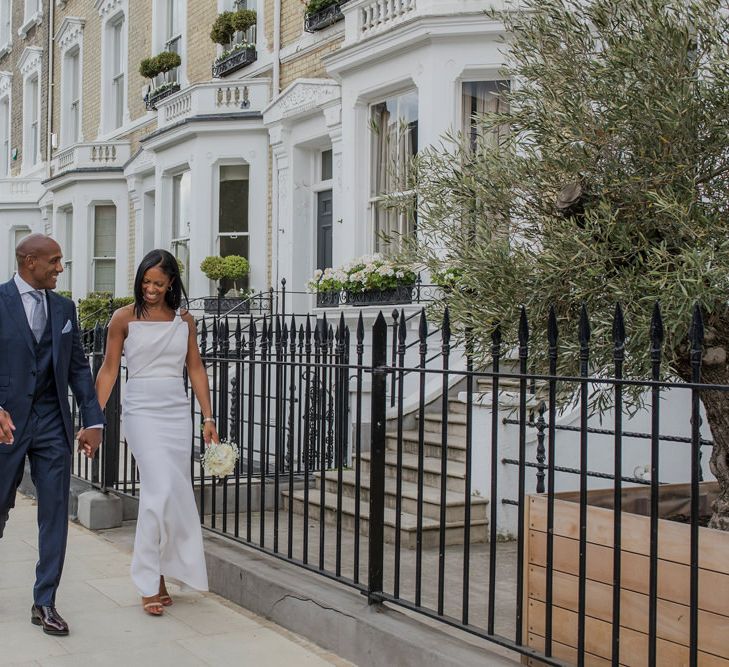 Stylish bride in a one shoulder fitted wedding dress holding hands with her groom in a navy Tom Ford Suit