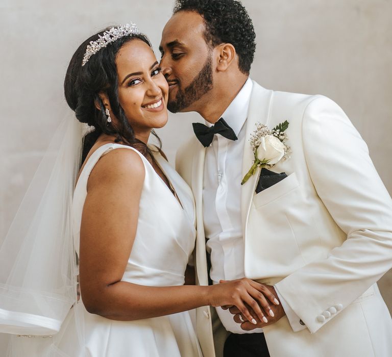 Groom in a white tuxedo jacket with white rose buttonhole flower kissing his brides cheek in a princess dress and tiara 