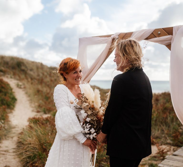 On bride in white dress faces other bride in black suit at boho elopement ceremony