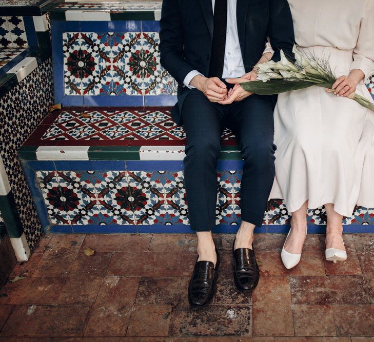 Couple sit on colourfully tiled bench, one holding bouquet and wearing cream dress, one in black suit and shoes with no socks