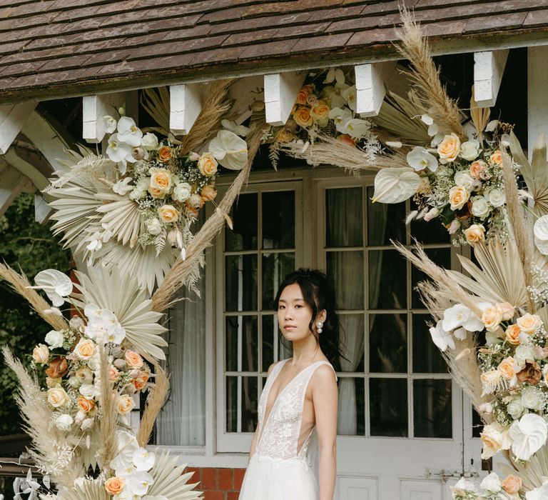 Bride at Berwick lodge wearing a plunge neck wedding gown with tulle skirt, under an arch of dried palms, pampas grass, white anthuriums, roses and orchids