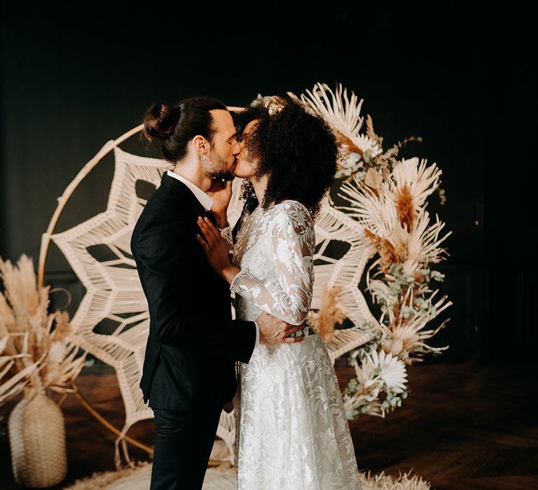 Bride & groom kiss in front of Macrame backdrop surrounded by dried florals