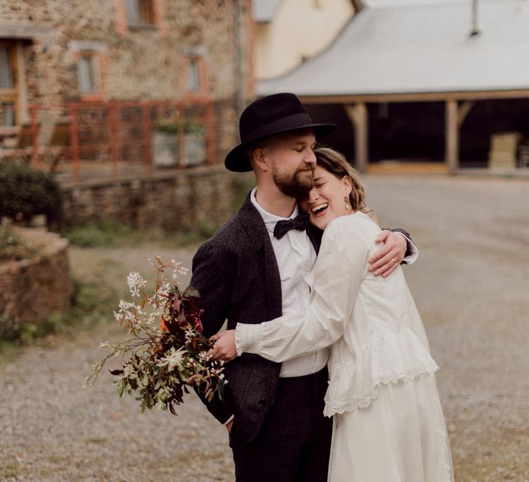 Laughing bride in white blouse, Charlie Brear wedding dress and applique veil holds multicoloured wedding bouquet whilst hugging groom in brown tweed suit, bow tie and black fedora at garden party wedding in Devon