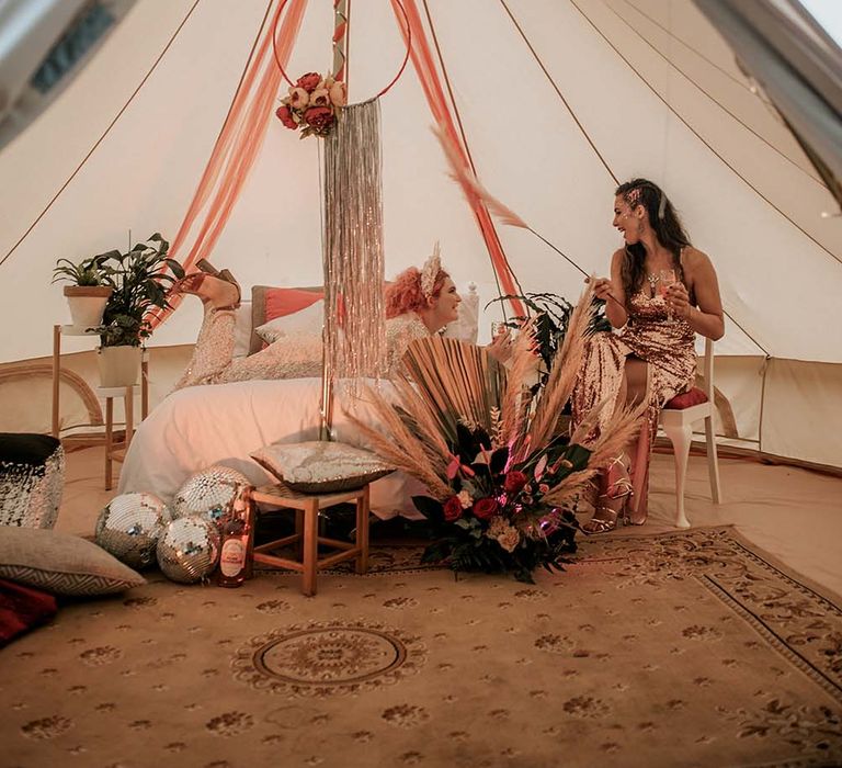 Two brides in sequin wedding dresses laughing together inside teepee with white bed, brown rug, disco balls and silver streamer decor for festival themed wedding