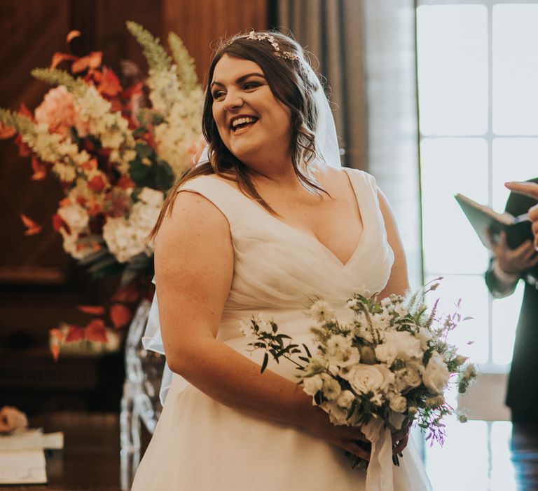 Bride smiles during wedding ceremony as she wears low neck wedding gown and holds white floral bouquet filled with roses and green foliage