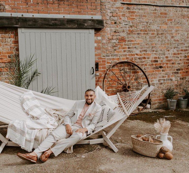 Groom in cream suit sitting on a large hammock for tropical wedding theme