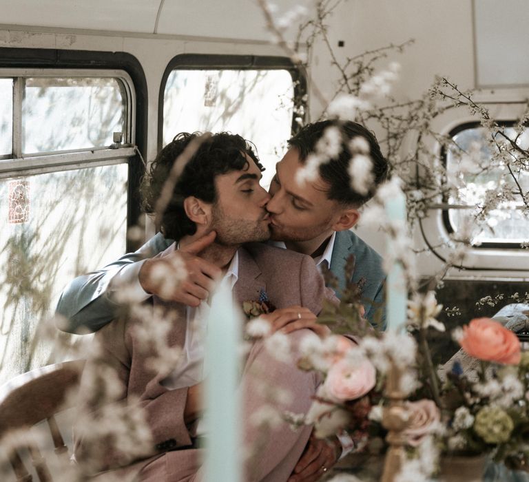 Grooms kiss whilst sat at rustic wooden table with pastel floral bouquets 