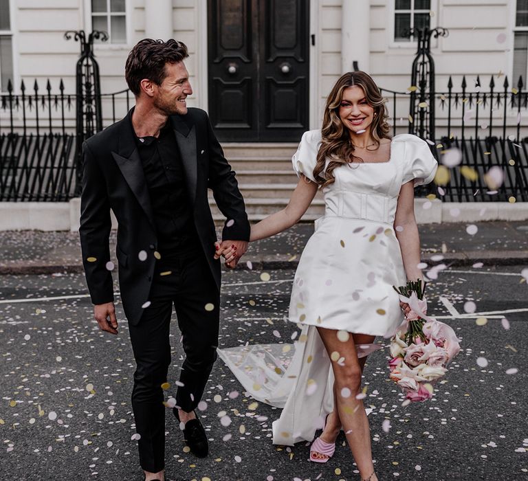 Confetti moment in the streets of London with bride in a knee length wedding dress with training bow back detail and groom in an all black suit 