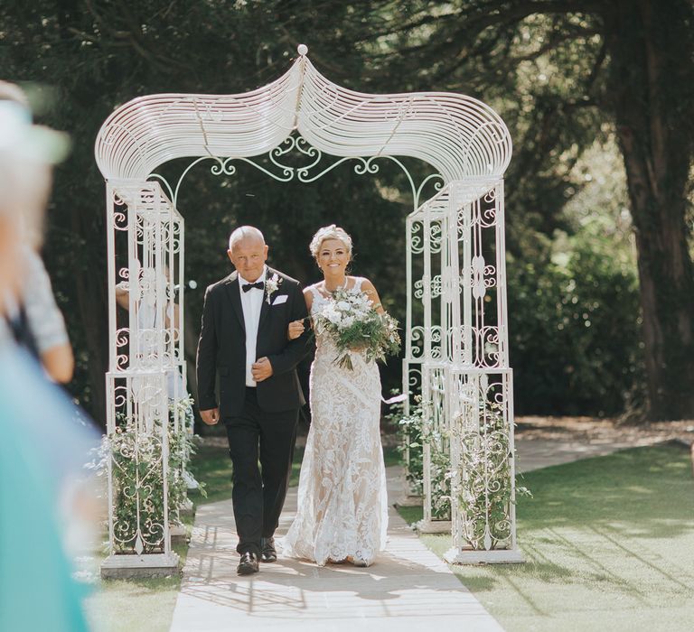 A bride in a lace wedding dress carries a bouquet and is linked arms with a man in a black tuxedo. They walk down an outdoor aisle.