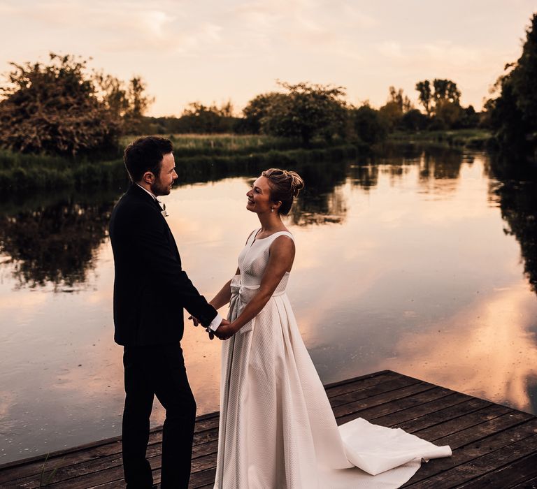 Bride & groom stand beside lake as the sun sets and they look lovingly at one another 