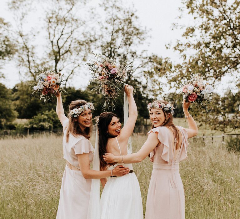 Bride in white cami wedding dress stands in between two bridesmaids in blush pink bridesmaids dresses as they hold up multicoloured wedding bouquets at garden wedding reception