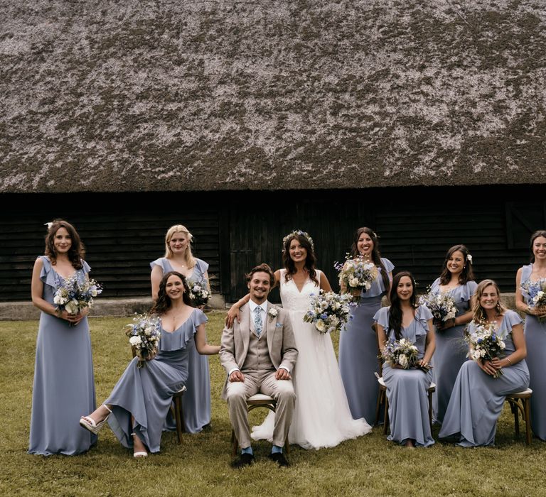 Bride & groom stand within fields for their post wedding pictures as bridesmaids wear dusty blue bridesmaids dresses and floral crowns