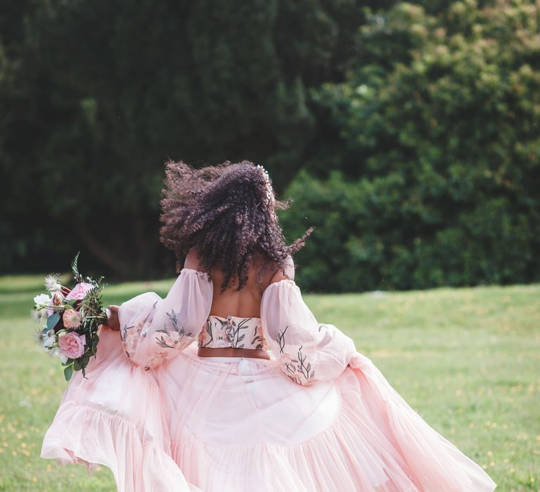 black bride with long naturally curly hair running in a garden in blush pink bridal separates with tulle skirt, embroidered bodice and detachable sleeves 