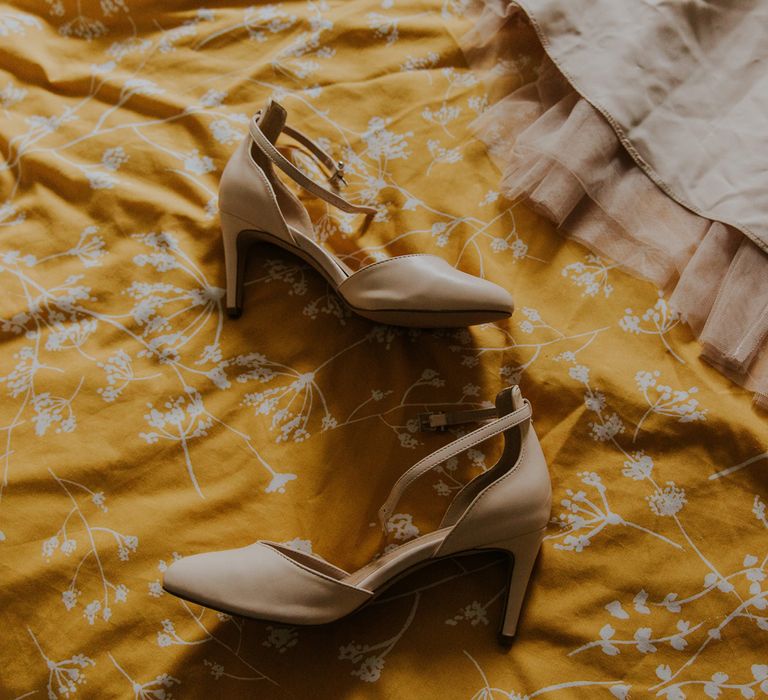 Brides pearl wedding dress and short nude strapped heels lay on yellow bedspread before barn wedding ceremony