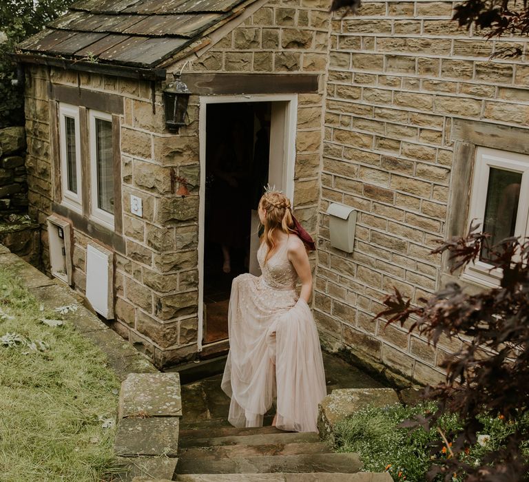 Bride in pearl wedding dress and crown holding red and green bridal bouquet leaves stone house before barn wedding ceremony