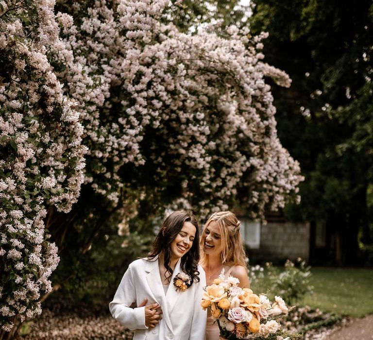 Bride in a blush pink tulle wedding dress holding a honey yellow and pink bouquet laughing with her bride in a white suit 