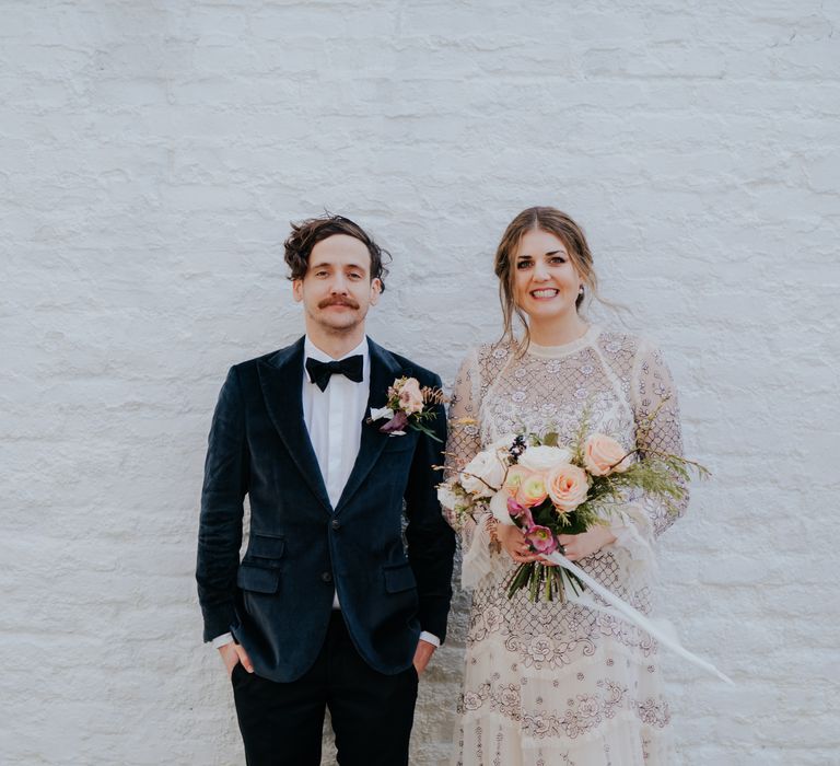 Bride & groom stand in front of white wall on their wedding day as groom wears deep velvet green suit with bow tie and bride wears Needle And Thread wedding dress