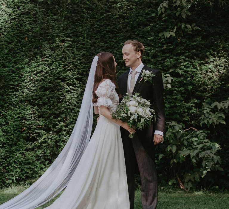 Bride in white lace puffed sleeve Daalarna wedding dress and bespoke veil holding white and green bridal bouquet stands looking at groom in black morning coat in the garden after church wedding ceremony in Surrey