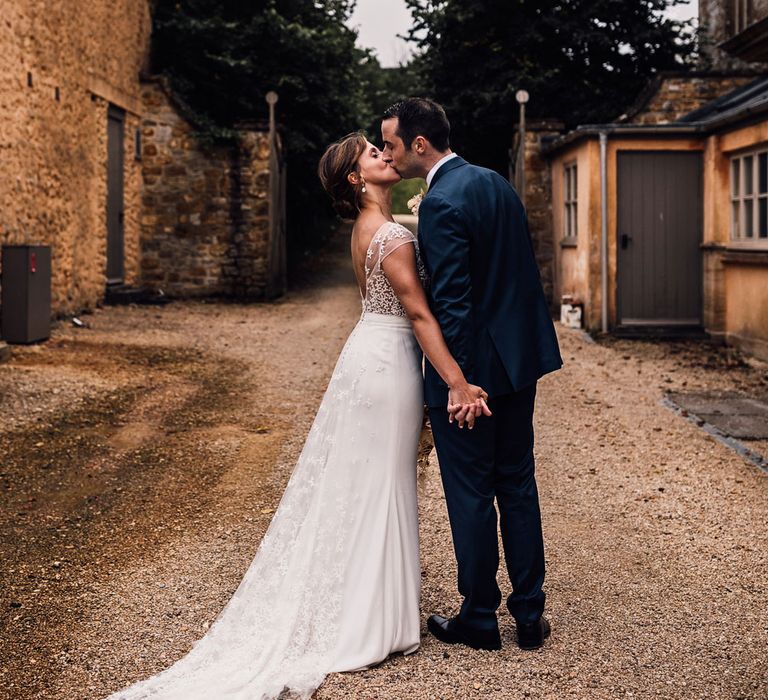 Bride in open back Rime Arodaky wedding dress with train holds hands with groom in blue suit as they kiss outside in courtyard at summer wedding in Dorset