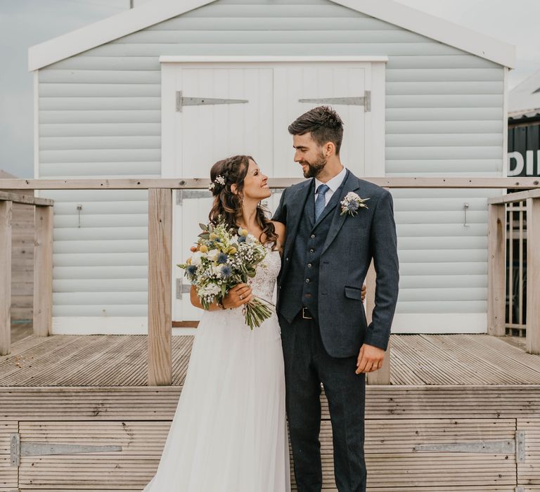 Bride & groom look lovingly at one another as they stand on the beachfront in front of hut on their wedding day