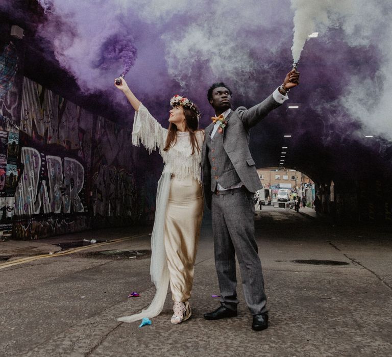 Black groom in a grey check suit holding a white smoke flare and a bride in a champagne gold satin wedding dress with tassels holding a purple smoke bomb