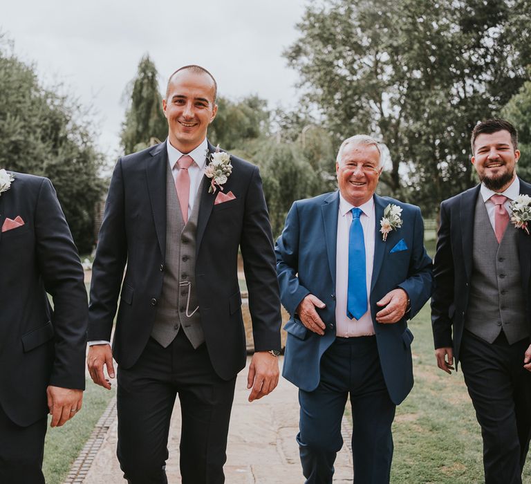 Groom and groomsmen in dark three piece suits with grey waistcoats, pink ties, pink pocket squares and protea buttonholes walking through gardens before Tythe Barn wedding with barn wedding flowers